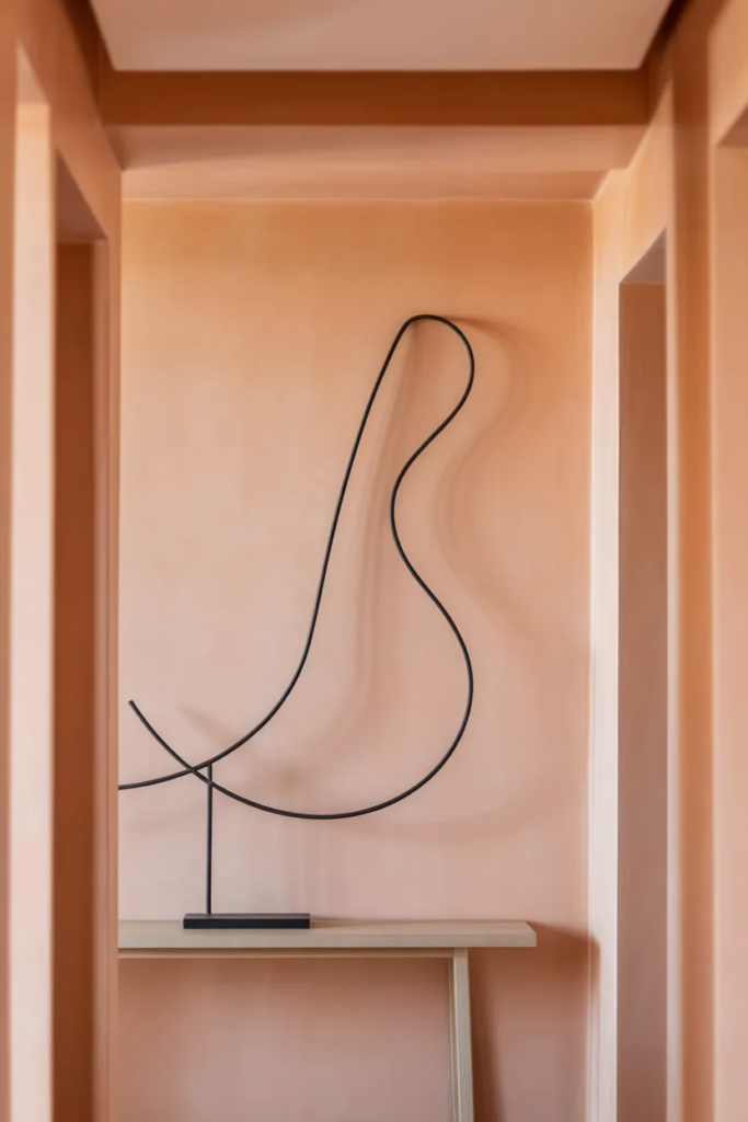 A minimalist black wire face sculpture mounted on a soft terracotta hallway wall. Dramatic side-lighting casting a sharp shadow, realistic mounting hardware, matte metal finish, 35mm lens, interior photography.