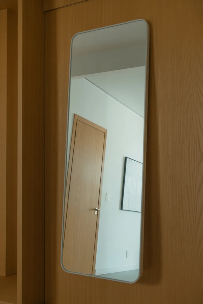 A tall, frameless vertical mirror leaning against a light oak hallway wall. Reflection of a minimalist interior, realistic glass smudges near the bottom, natural morning light, wide-angle lens, interior photography.