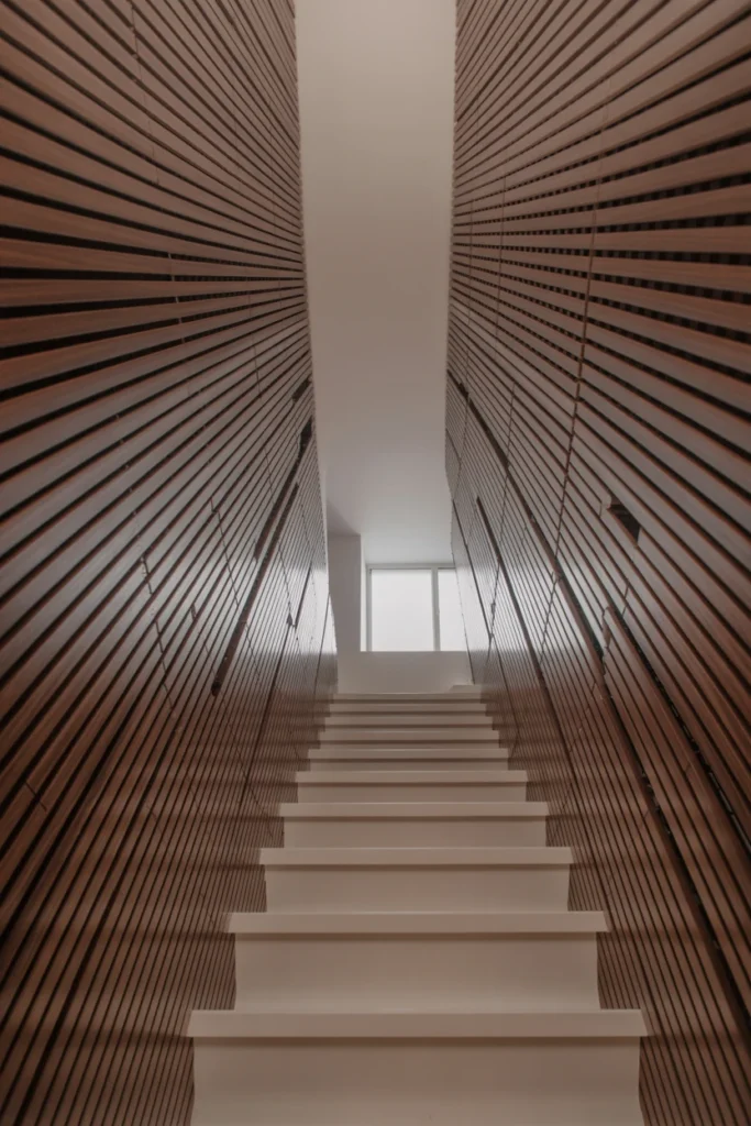 An ultra-wide 14mm architectural shot taken from the bottom of a narrow staircase looking up. The side wall is covered in thin, vertical walnut wood fins spaced 4 inches apart. A 14mm lens captures the dramatic perspective of the stairs and the high ceiling. Soft natural light from a window at the top of the stairs creates rhythmic shadows across the fins. High-fidelity wood grain and realistic plaster texture. Professional interior photography.