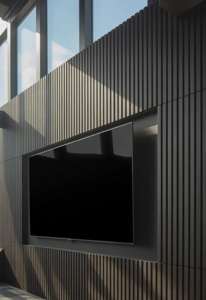 An eye-level, medium-wide shot of a media room. The entire wall is covered in matte charcoal-stained vertical wood slats. A large TV is mounted in the center. Strong side-lighting from a large window during midday highlights the deep shadows between the slats and the gritty wood texture. 50mm lens, high-contrast architectural photography.