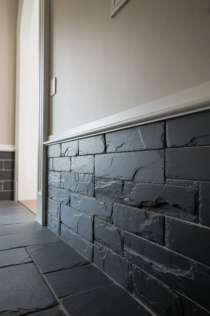 An ultra-wide 14mm architectural shot of a farmhouse entryway. The lower half of the wall is clad in dark, jagged slate tiles with deep grout lines. The 14mm lens captures the transition from the stone wainscoting to the white wall above. Soft light glints off the "oily" surface of the stone, revealing its realistic riven texture. Professional interior photography.