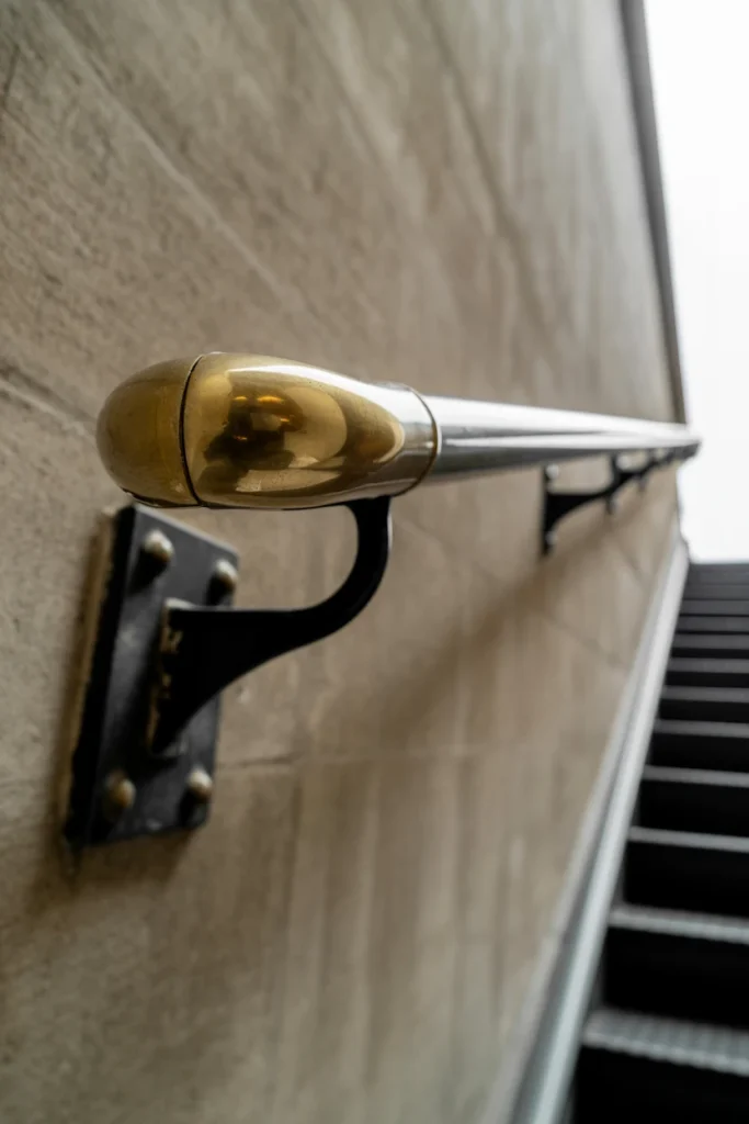 A wide-angle 20mm shot looking down a narrow staircase. A sleek, solid brass handrail runs the length of the wall, held by heavy industrial black mounts. The lighting is "Overcast Day," providing even illumination that shows the realistic mottled tarnish and bright polished sections of the brass. The lens captures the perspective of the stairs narrowing into the distance. Professional interior photography.
