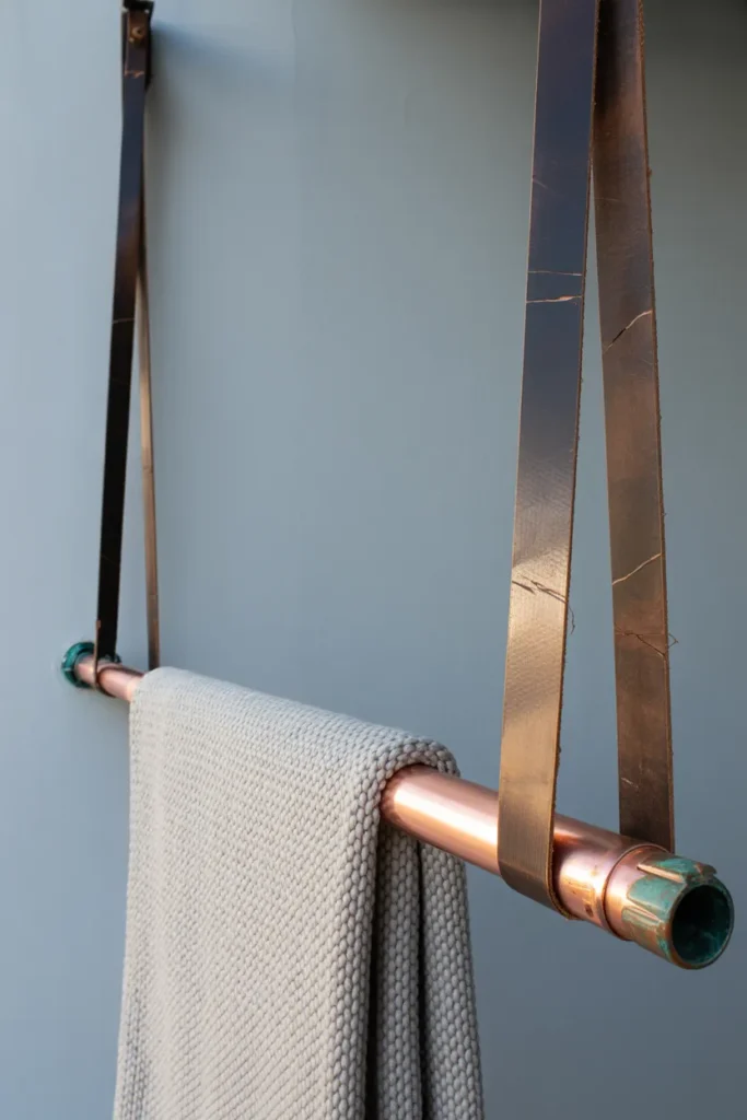A horizontal copper pipe hung by brown leather straps on a light grey wall. A folded blanket hanging over it, realistic metal patina, natural morning light, 35mm lens.