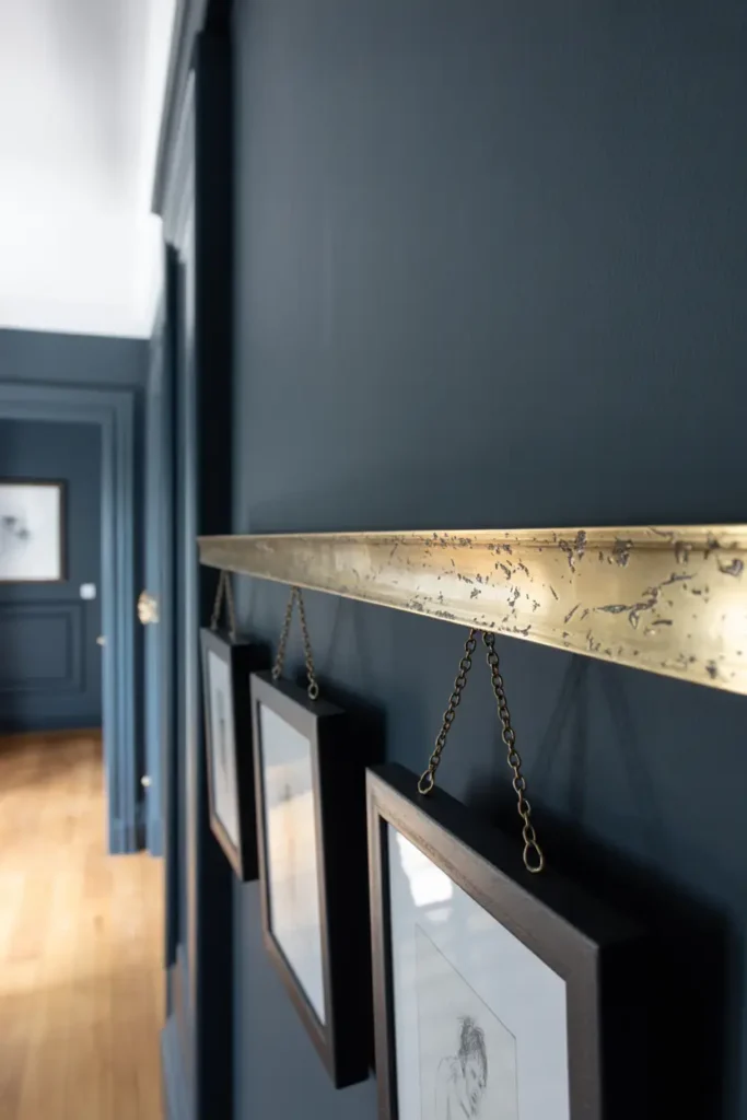 A thin aged brass picture rail on a deep charcoal wall. Minimalist frames hanging from delicate chains, soft side-lighting, realistic metal tarnish, interior design photography, high-detail texture.