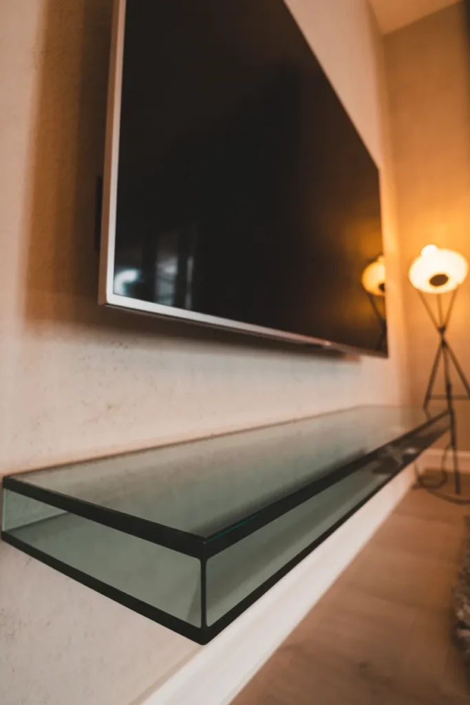 A wide-angle, low-shot of a modern living room at night. A large TV is mounted on a white plaster wall with a thick, smoked-grey glass ledge floating beneath it. A single warm floor lamp in the corner reflects softly off the glass surface. Realistic glass edges and a few faint fingerprints visible. 35mm lens, moody and atmospheric interior photography.
