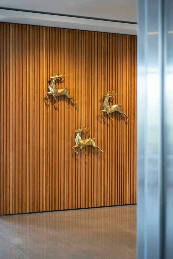 An ultra-wide B2B style shot of a modern hallway feature wall. The wall is covered in floor-to-ceiling vertical oak timber slats. Crisp, polished brass floating geometric deer silhouettes are mounted perfectly across the wood paneling using architectural standoffs. Sophisticated, bright daylight, professional architectural styling.