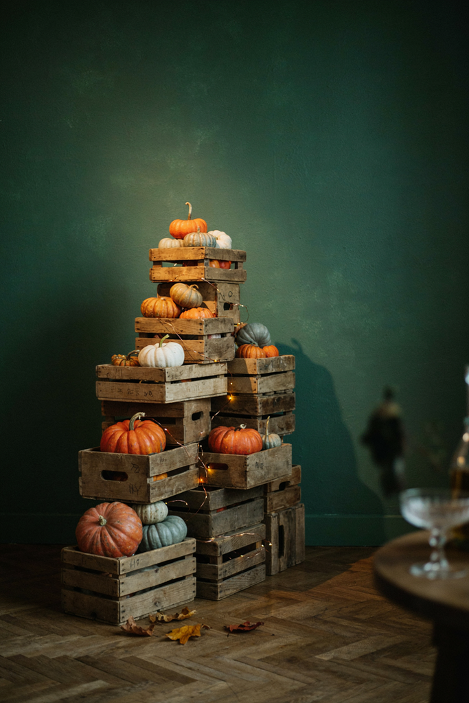 Stacked wooden crates holding orange and white pumpkins against a green wall with fairy lights