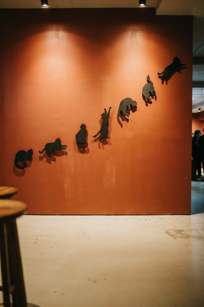 Seven black cat silhouettes in different poses walking across a burnt orange wall