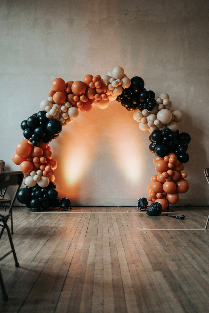 Black, orange, and cream balloon garland arch on a bone wall lit by two floor uplights