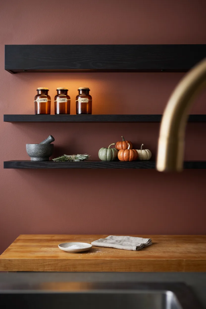 Two black oak shelves with amber glass bottles and mini pumpkins on an oxblood kitchen wall