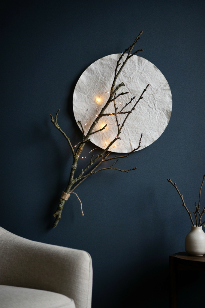 Textured white paper moon behind bare branches wrapped in warm string lights on a dark navy wall
